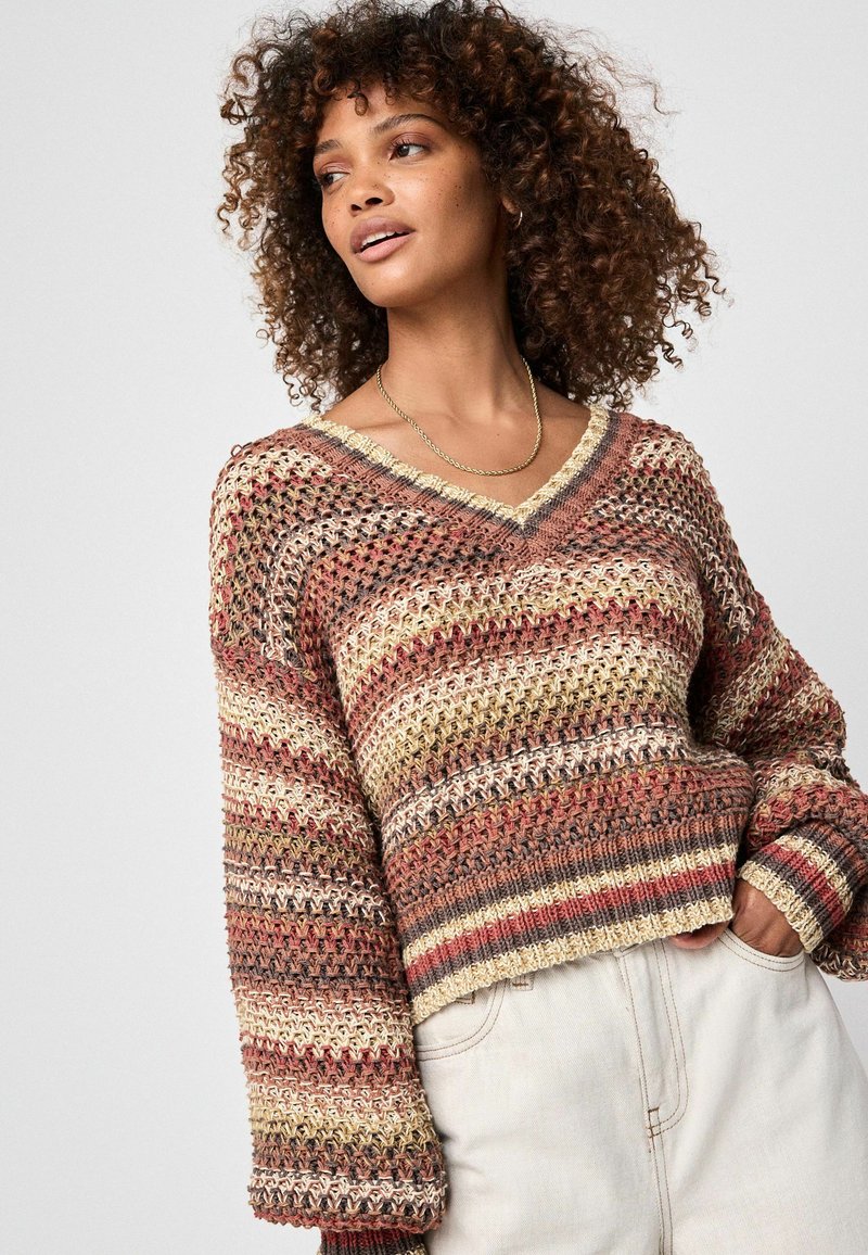 Femeie îmbrăcată într-un pulover tricotat în culori multiple, cu decolteu în V și mâneci lungi, purtând pantaloni deschiși la culoare, cu talie înaltă, cu mâna în buzunar.