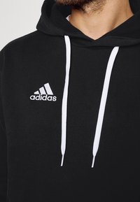 Sweat à capuche noir Adidas avec cordons blancs porté par une personne, avec le logo Adidas sur la poitrine gauche.