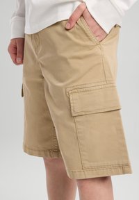 Beige cargo shorts made of cotton, featuring side pockets, stitched detailing, and a relaxed fit. The hem is slightly rolled.