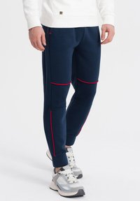 Marinblå joggerbyxor med röda accentstråk, ribbad textur och resårmidja. Matchas med ljusgrå sportskor.