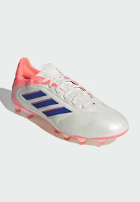 Zapatilla deportiva blanca de fútbol con acabado brillante, acentos en rosa, rayas moradas, parte superior texturizada y tacos transparentes para tracción.