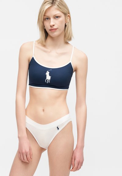 Femme portant un haut court bleu marine avec bordure blanche et un slip blanc, les deux ornés d'un logo de joueur de polo, posant devant un fond uni.