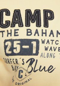 Camiseta de algodón amarilla con texto bordado azul. Presenta frases como "CAMPAMENTO EN LAS BAHAMAS" y "LA BAHÍA DEL SURFISTA", añadiendo interés visual a través de la tipografía.