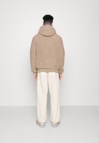Sudadera con capucha de forro polar beige con una capucha grande y suave y puños acanalados, combinada con pantalones anchos de pierna ancha de color blanco roto y calzado claro. Fondo liso.