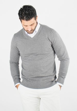 Un pull gris en V fabriqué à partir d'un matériau tricot doux, présentant des manches longues et des poignets côtelés, porté par-dessus une chemise blanche à col.