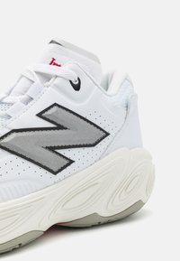 Zapatilla deportiva blanca con un upper texturizado y logo plateado. Suela ondulada distintiva con un toque de rojo en el talón. Presencia de acentos en negro.