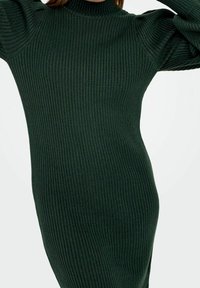 Femme portant une robe pull moulante à col roulé côtelé, vert foncé, à manches longues, bras levés près de sa tête.