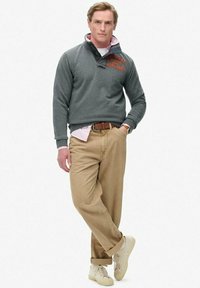 Sudadera de forro polar gris con cuello abotonado y logotipo naranja; llevada sobre una camisa rosa clara. Pantalones beige y zapatillas crema completan el atuendo.