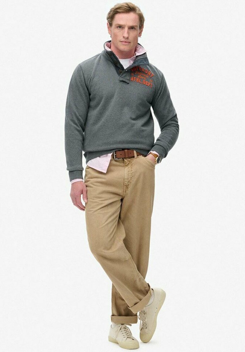 Sudadera de forro polar gris con cuello abotonado y logotipo naranja; llevada sobre una camisa rosa clara. Pantalones beige y zapatillas crema completan el atuendo.