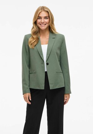 Olivově zelená blazer s jedním knoflíkem na zapínání, klopovými límci, dvěma předními kapsami a přiléhavým střihem. Nosené přes bílé tričko a černé kalhoty.