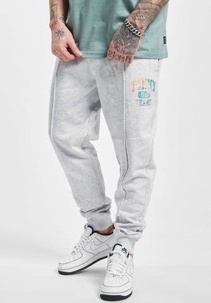 Man wearing light gray sweatpants with colorful "Tommy Jeans" logo, white Nike sneakers, and a green shirt, showing tattooed hands.