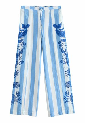 Vrouw die een blauw mouwloos topje draagt, brede gestreepte broek met zeeleven prints en een strooien hoed, staand bij zee met rotsen en golven.