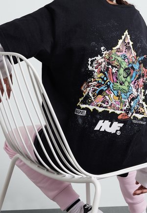 Osoba siedzi na białym drucianym krześle, nosi czarną koszulkę z grafiką superbohaterów Marvela z Thorem, Hulkiem, Iron Manem i innymi, różowe spodnie i buty Adidas.