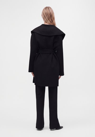 Femme aux longs cheveux blonds vue de dos, portant un manteau noir ceinturé avec un large col et un pantalon noir sur un fond clair uni.