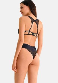 Ensemble de lingerie noire comprenant un bralette en dentelle avec des bretelles réglables et un string assorti. Le bralette présente un détail en maille au dos.