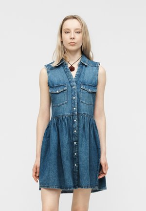 Ung kvinde iført en ærmeløs blå denimskjortekjole med brystlommer foran og en halskæde med en rød blomst vedhæng.