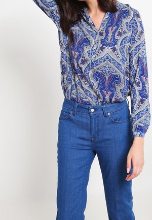 Woman wearing blue paisley-patterned blouse tucked into high-waisted blue denim jeans, one arm raised above her head.