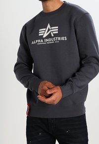Sweatshirt gris foncé avec un logo blanc et le texte "ALPHA INDUSTRIES". Manches et ourlet côtelés, avec un détail de tag rouge sur le côté.