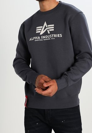 Sweatshirt gris foncé avec un logo blanc et le texte "ALPHA INDUSTRIES". Manches et ourlet côtelés, avec un détail de tag rouge sur le côté.