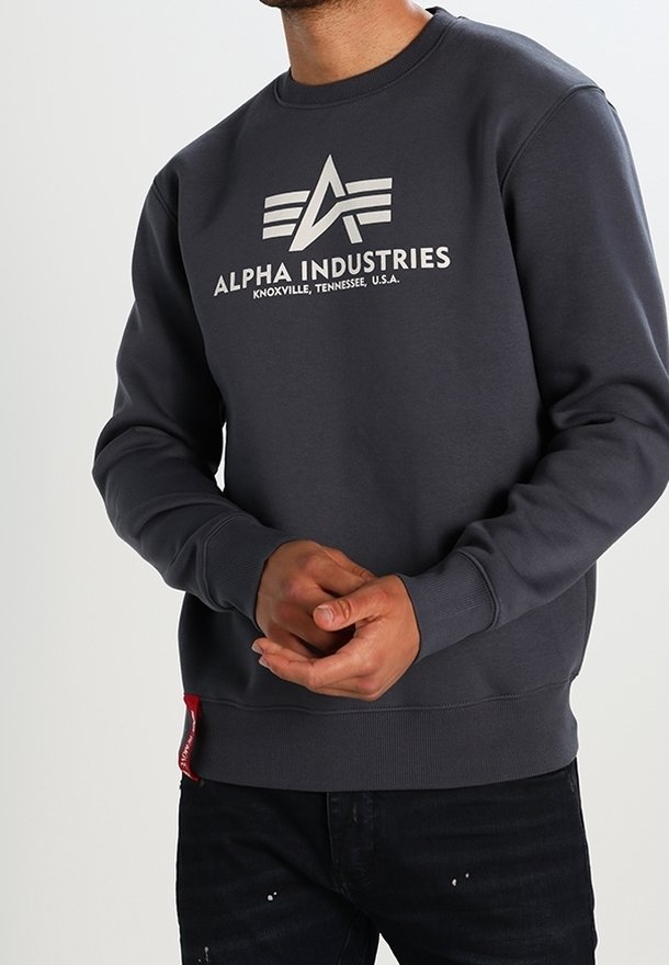 Sweatshirt gris foncé avec un logo blanc et le texte "ALPHA INDUSTRIES". Manches et ourlet côtelés, avec un détail de tag rouge sur le côté.
