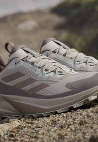 Para beżowo-brązowych butów do biegania w terenie Adidas Terrex na kamienistym podłożu z teksturowaną podeszwą i zawiązanymi sznurówkami.