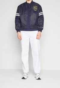 Námořnická bomber bunda s logem, bílé pruhy na rukávech, zip vpředu. Společně s bílými kalhotami a šedými teniskami.