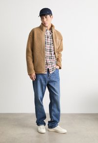 Brun fleecejakke med krave, rutet skjorte under, blå jeans og hvide sneakers. En marineblå kasket tilføjer et kontrasterende accent.