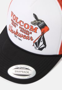 Kravas automašīnu cepure ar baltu augšu, sarkaniem sietiem un melnu vizieri. Satur Volcom zīmola logo un grafiku ar otu. Ar snapback aizdari.
