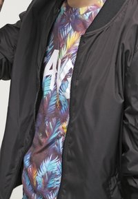 Black bomber jacket with a smooth, shiny texture, worn over a colorful, patterned tropical print T-shirt featuring large white lettering.