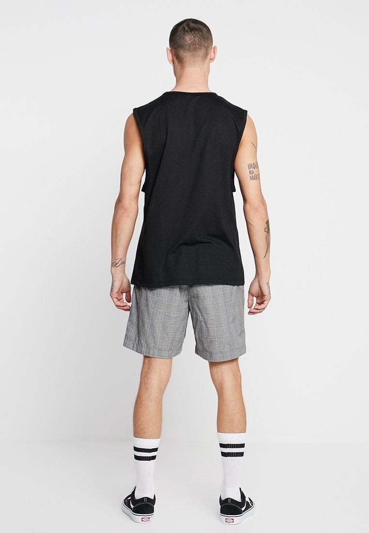 Chemise sans manches noire en coton, associée à un short à carreaux gris, des chaussettes blanches avec des rayures noires et des chaussures noires.