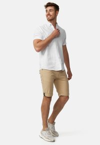 Vit kortärmad skjorta med knappar, bruna uppvikta shorts med svarta detaljer, och ljusgrå sneakers. Enkel, avslappnad och åtsittande design.