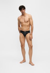 Jeune homme aux cheveux bouclés foncés portant un slip noir à motifs, se tenant devant un fond clair uni.