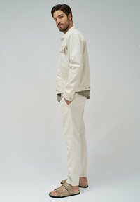 Chaqueta de mezclilla crema, camisa oliva y joggers beige. Sandalias en tonos tierra con dos hebillas. Diseño sencillo y moderno con un ajuste relajado.