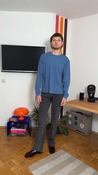 Mężczyzna w niebieskiej koszuli z długim rękawem i szarych dżinsach stoi w pokoju z telewizorem na ścianie, pomarańczową lampą, roślinami oraz stolikiem z manekinem głowy i walizką.