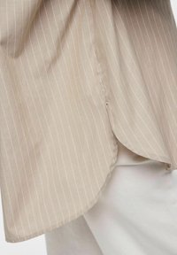 Camicia beige con righe verticali bianche, realizzata in tessuto morbido. Presenta un orlo curvo e una fessura laterale per facilitare il movimento.