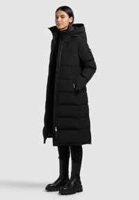 Schwarze Daunenjacke mit langem, gestepptem Design, Kapuze und Reißverschluss-Taschen; kombiniert mit schwarzen Knöchelstiefeln und eng anliegenden schwarzen Hosen.