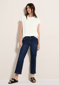 Haut blanc à manches courtes, coupe décontractée, associé à un jean cropped bleu foncé. Des sandales noires avec des accents dorés complètent la tenue. Design simple.