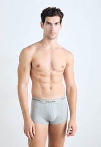 Calvin Klein sivi bombažasti bokserji s črnimi hlačnicami in logotipom, prilegajoč dizajn in gladka tekstura. Model stoji pokončno.