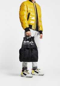 Persona che indossa una giacca a vento gialla, pantaloni grigio chiaro, scarpe da ginnastica bianche con dettagli gialli e neri, che tiene uno zaino Diesel nero per la maniglia superiore.