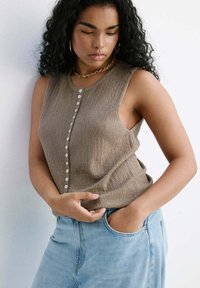 Femme aux cheveux bouclés porte un haut beige sans manches boutonné et un jean bleu clair, debout contre un mur blanc, une main dans la poche.