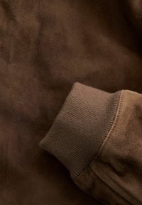 Tela de ante marrón con un puño de punto acanalado en un tono a juego, que muestra una textura suave y detalles de construcción sin costuras.