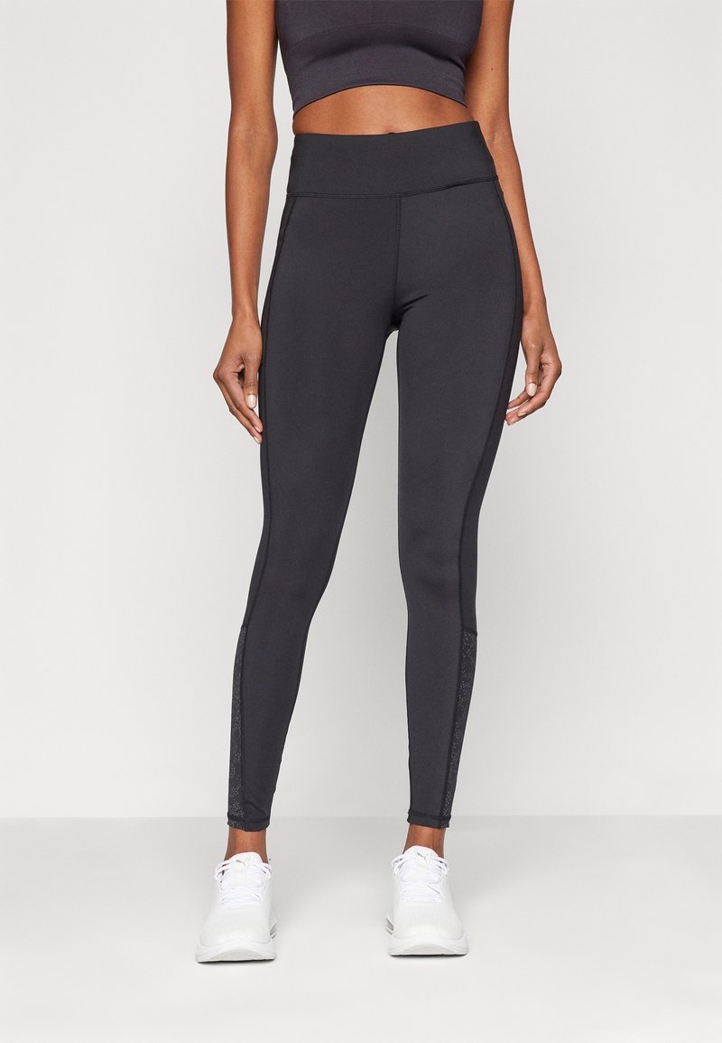 Leggings noirs avec une taille haute, présentant des panneaux latéraux texturés et une coupe ajustée. Associés à des chaussures de sport blanches.