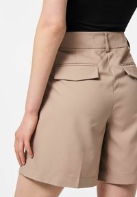 Shorts beiges sur mesure avec poches arrière. Le tissu semble lisse avec un design structuré, présentant un ourlet propre et une taille haute.