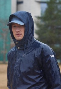 Mężczyzna w mokrej granatowej kurtce przeciwdeszczowej z kapturem i jasnoniebieską czapką, stojący na zewnątrz w deszczowy dzień.