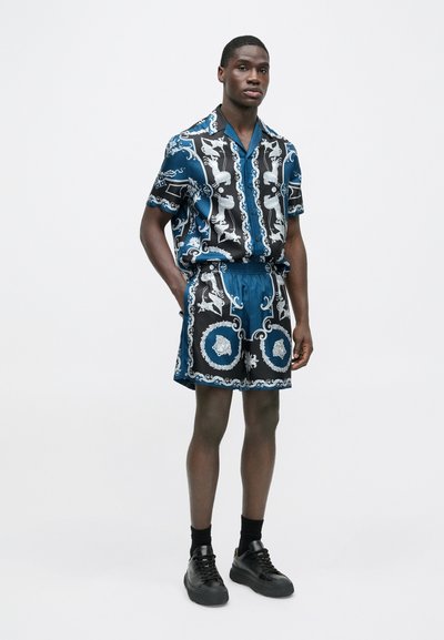 Košile s krátkým rukávem na knoflíky a kraťasy s elastickým pasem v tmavě modré barvě, zdobené bílými vzory. Model nosí černé tenisky a ponožky.