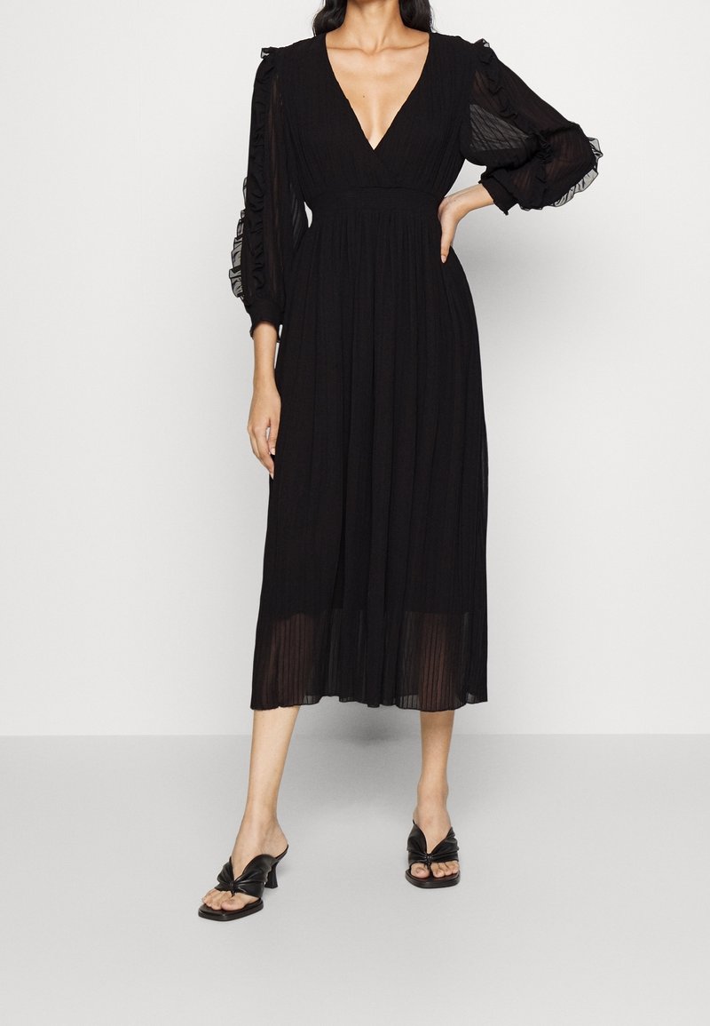 Femme portant une robe midi noire avec de longues manches bouffantes transparentes, un décolleté en V profond, une jupe plissée et des sandales à talons noires, debout avec une main sur la hanche.