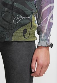 Blusa trasparente multicolore con motivi floreali, vita arricciata e polsini elasticizzati. Presenta un logo ricamato in bianco sul tessuto.