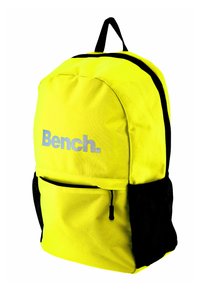 Żółony plecak z tkaniny, z czarną siatkową kieszenią boczną oraz przednią kieszenią na zamek. Na przedniej części znajduje się srebrne logo "Bench.".