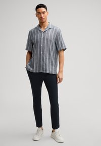 Korte mouwen overhemd in het grijs met witte verticale strepen, gecombineerd met navy slim-fit broek en witte sneakers. Effen achtergrond.