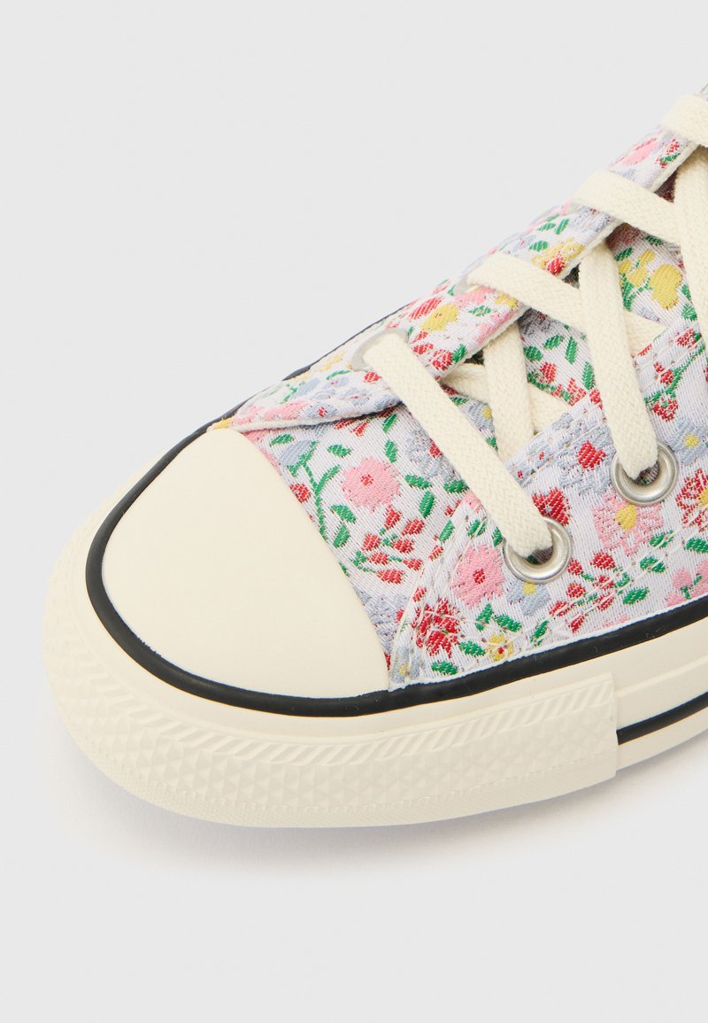 Leinwand-Sneaker mit Blumenmuster, beiger Gummispitze, beigen Schnürsenkeln und schwarzem Besatz an der strukturierten Sohle vor weißem Hintergrund.
